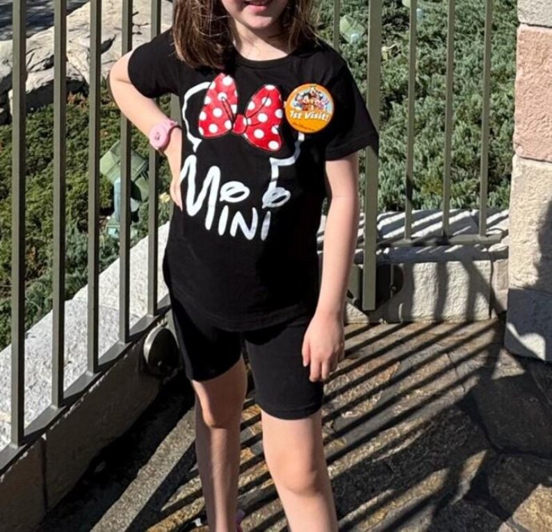 May include: A black short-sleeved t-shirt featuring a white outline of a bow and the text "MINI" in white. A red bow with white polka dots is positioned above the text. A round yellow "1st Visit" button is pinned to the upper chest area.