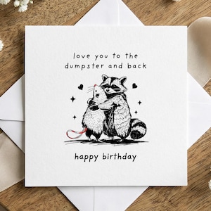 Puede incluir: Tarjeta de felicitación blanca con una ilustración en blanco y negro de un mapache abrazando a una rata. La tarjeta dice "love you to the dumpster and back" y "happy birthday". La tarjeta está sobre una superficie de madera.