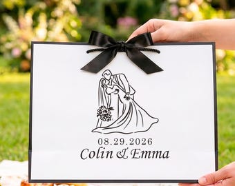 Puede incluir: Una bolsa de regalo rectangular blanca con bordes negros y un lazo de cinta de satén negro en la parte superior. El frente presenta una ilustración de línea negra de una pareja besándose, la fecha 29.08.2026 y los nombres Colin & Emma.