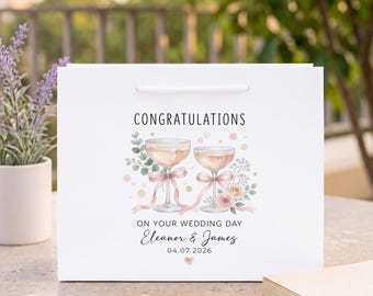 Puede incluir: Una bolsa de regalo blanca con el texto CONGRATULATIONS ON YOUR WEDDING DAY, Eleanor & James, y 04.07.2026. El diseño incluye dos copas de champán con lazos rosas, detalles florales y un pequeño icono de corazón en tonos pastel suaves.