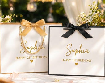 Puede incluir: Dos bolsas de regalo blancas con detalles en oro y negro. Cada bolsa presenta el nombre Sophia en letra dorada, el texto HAPPY 21ST BIRTHDAY y un pequeño corazón dorado. Una bolsa tiene un lazo dorado y asas de cuerda, la otra un lazo negro.