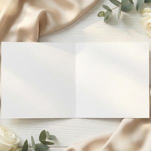 Puede incluir: Una tarjeta de felicitaci&oacute;n abierta y en blanco con interior blanco, sobre una superficie de madera blanca. La tarjeta est&aacute; rodeada de tela de seda beige, rosas blancas y hojas de eucalipto verdes, creando una est&eacute;tica elegante y natural.