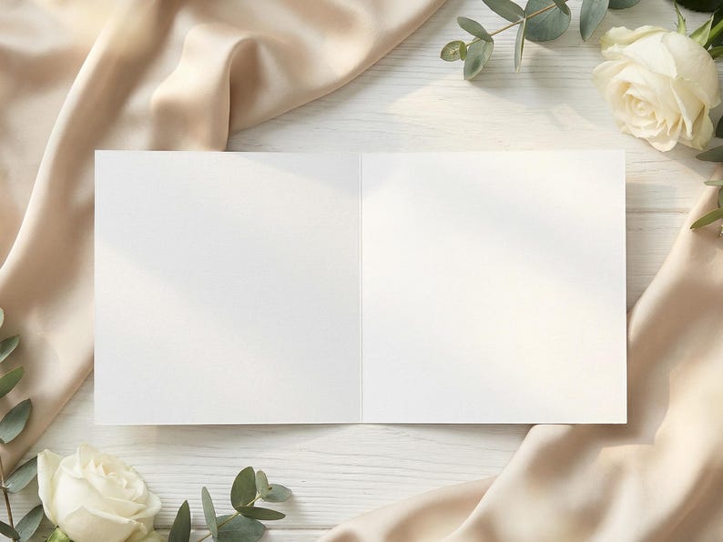 Puede incluir: Una tarjeta de felicitaci&oacute;n abierta y en blanco con interior blanco, sobre una superficie de madera blanca. La tarjeta est&aacute; rodeada de tela de seda beige, rosas blancas y hojas de eucalipto verdes, creando una est&eacute;tica elegante y natural.