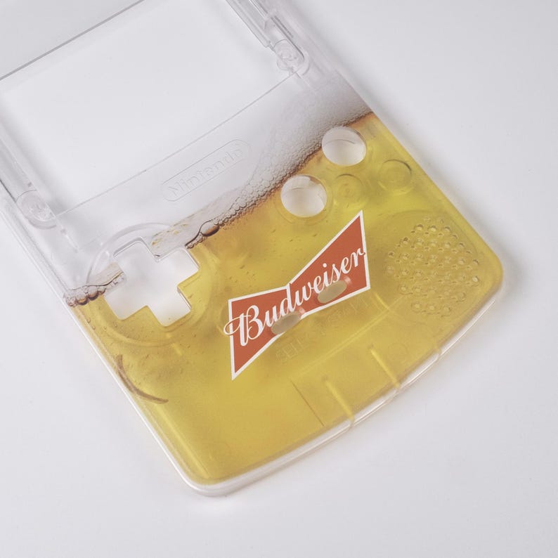 May include: Clear plastic Game Boy case with a beer-themed design. The case features a yellow and foamy beer graphic with the Budweiser logo. The Nintendo logo is visible on the top of the case.