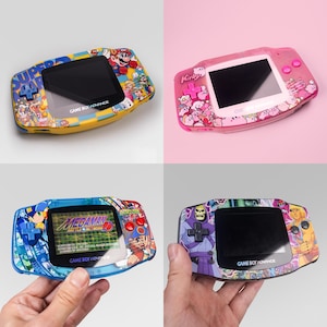 Op de afbeelding: Vier Game Boy Advance handheld consoles met aangepaste behuizingen met karakterafbeeldingen. Eén is geel met Super Mario, één is roze met Kirby, één is blauw met Mega Man en één is paars met He-Man en Skeletor karakterillustraties.