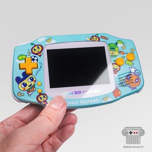 May include: A light blue Game Boy Advance handheld console with a white screen and colourful cartoon character designs. The console features a directional pad, action buttons, and the text "GAME BOY ADVANCE" and "NGC SCHOOL".