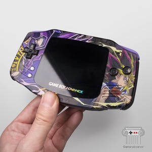 May include: A Game Boy Advance handheld console with a custom purple and black design featuring anime characters. The console has a black screen and buttons, with the text "GAME BOY ADVANCE" printed on the front.