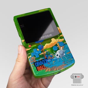 Op de afbeelding: Een groene Game Boy Color handheld console met een Dragon Ball-ontwerp. De console heeft een zwart scherm, een D-pad en actieknoppen. Het Dragon Ball-ontwerp bevat personages en landschappen in blauw, oranje en groen.