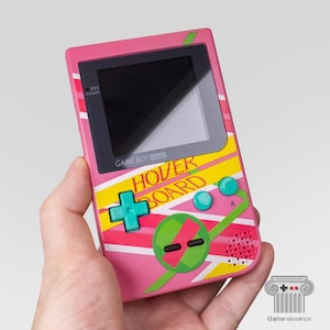 May include: A pink handheld video game console featuring a screen, a directional pad, and two buttons. The device is decorated with yellow, white, and green diagonal stripes and the text HOVER BOARD printed in red letters across the front surface.
