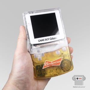 May include: A clear Game Boy Color handheld console filled with beer, featuring a Budweiser logo. The screen is black, and the text "GAME BOY COLOR" is visible. The device is held in a hand.