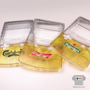 May include: Three clear Game Boy Color cases filled with beer, each featuring a different brand logo: Carlsberg, Budweiser, and Heineken. The cases are partially filled with a golden liquid, resembling beer, and are angled slightly.