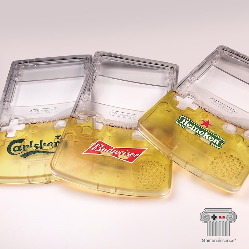 May include: Three clear Game Boy Color cases filled with beer, each featuring a different brand logo: Carlsberg, Budweiser, and Heineken. The cases are partially filled with a golden liquid, resembling beer, and are angled slightly.