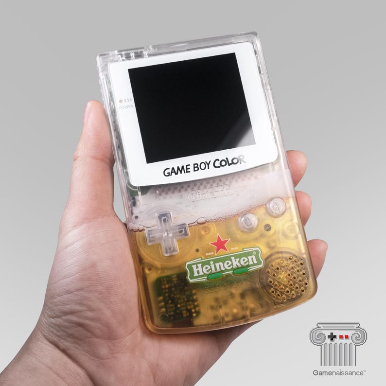 May include: A clear Game Boy Color handheld console with a Heineken beer design. The console has a black screen, white border, and the words "GAME BOY COLOR" printed on it. The Heineken logo is visible on the lower part of the console.