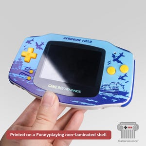Op de afbeelding: Een handheld Game Boy Advance console met een aangepast ontwerp. De behuizing is lichtblauw met een afbeelding van vliegtuigen en oceaangolven. De knoppen zijn geel. De tekst "AEROGUN FIELD" en "GAME BOY ADVANCE" zijn zichtbaar.