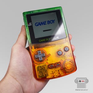 Peut inclure: Une console de jeu portable Game Boy Color avec une coque transparente dégradée orange et verte. L'appareil dispose d'un écran affichant le texte GAME BOY, d'une croix directionnelle, de boutons A et B, ainsi que de boutons start et select gris.