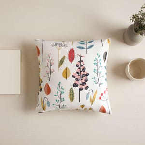 May include: A square decorative pillow with a white background and a colorful botanical pattern featuring leaves and flowers. A small vase with greenery, a mug, and a blank book are also on the table.