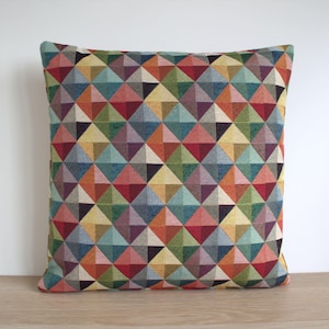Op de afbeelding: Vierkant kussen met een geometrisch patroon van veelkleurige driehoeken in tinten rood, oranje, geel, groen, blauw en paars. Het kussen is gemaakt van een textuurstof, geschikt voor interieurdecoratie.