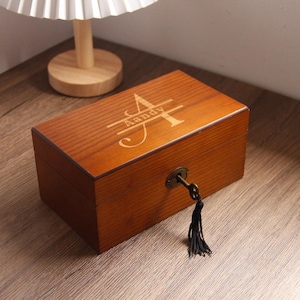 May include: A rectangular wooden box with a lock and key, featuring the name "Aandy" engraved on the lid. The box is a warm brown colour and has a black tassel attached to the key. The box is sitting on a wooden surface.