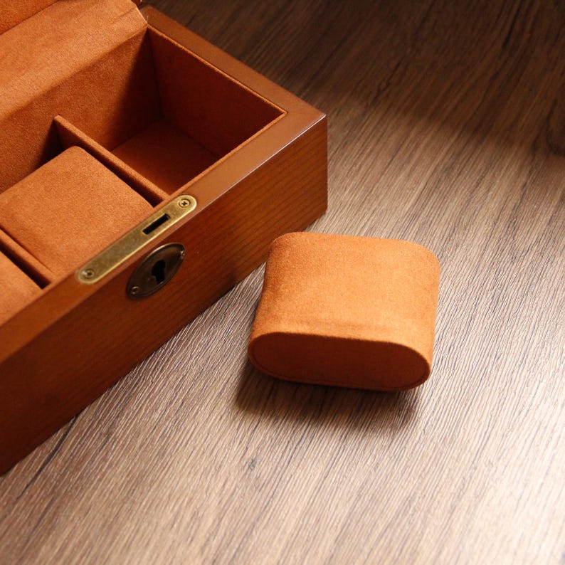 May include: A wooden watch box with a brass clasp and keyhole, lined with orange felt. An orange felt watch pillow sits beside the open box. The box is on a wooden surface.