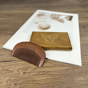 Op de afbeelding: Een houten kam en een houten doos met een vlinderontwerp en de naam "Emily" gegraveerd. De kam is donkerbruin met fijne tanden. De rechthoekige doos heeft een scharnierend deksel. De achtergrond omvat wit papier en een houten oppervlak.