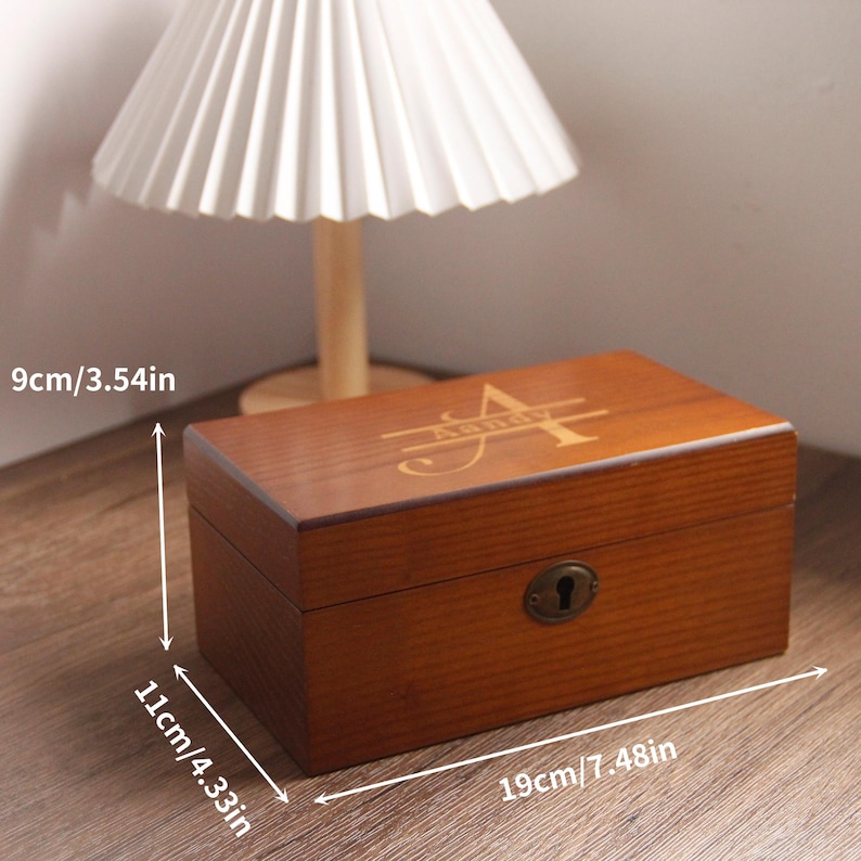 May include: A brown wooden box with a lock and an initial design on the lid. The box measures 19cm long, 11cm high, and 9cm deep. The box has a vintage aesthetic.