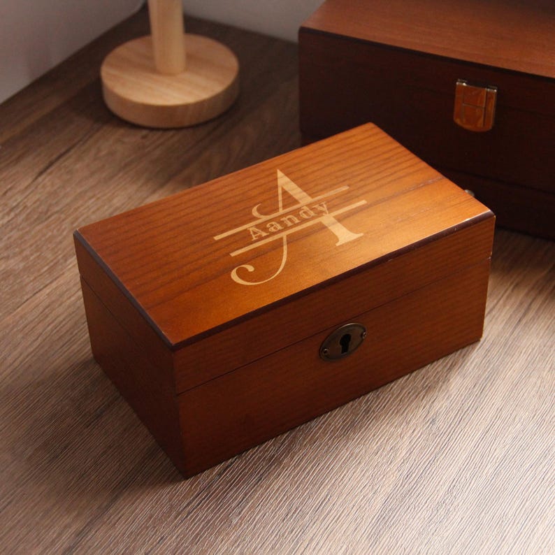 May include: A rectangular wooden box with a lock and keyhole. The box is a warm brown colour and has the name "Aandy" engraved on the lid in a decorative font. Another wooden box is visible in the background.
