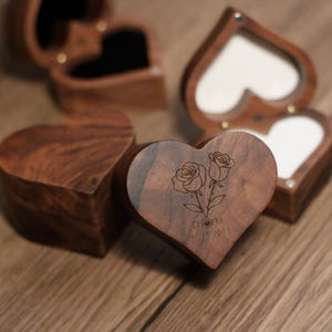 May include: Heart-shaped wooden ring boxes with a dark brown finish. The boxes feature a rose design and the initials "D M" with the date "02 14 2003" engraved on the lid. Some boxes are open, revealing a white interior.