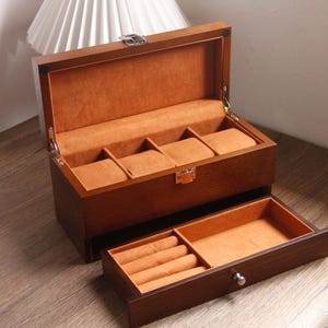 May include: A wooden watch box with a hinged lid and a pull-out drawer. The box is a warm brown colour with a soft orange interior. The top section has compartments for watches, and the drawer has ring storage.