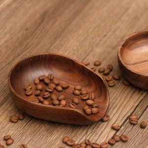 Puede incluir: Un cuenco de madera en forma de cuchara lleno de granos de café, junto a un cuenco redondo de madera. Los cuencos y los granos son de color marrón cálido, colocados sobre una superficie de madera. La cuchara tiene una extensión en forma de asa.