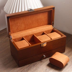 May include: A dark brown wooden watch box with a hinged lid and a drawer. The interior is lined with orange-brown fabric and has three watch pillows. A separate watch pillow is visible. The box is designed for watch storage.