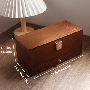 May include: A rectangular wooden box with a drawer, measuring 23.5 cm long, 10.5 cm wide, and 11.5 cm high. The box has a dark brown finish, a metal clasp, and a small drawer pull. The box is sitting on a wooden surface.