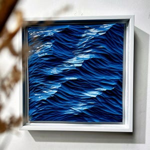 May include: A square wall art piece featuring a deep blue, textured relief sculpture that mimics the appearance of ocean waves. The artwork is encased in a simple, white wooden frame, creating a high-contrast visual effect with the dark blue tones.