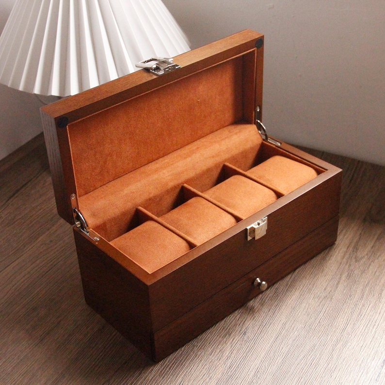 May include: A dark brown wooden watch box with a hinged lid, open to show four watch compartments. The interior is lined with orange-brown fabric. The box has a silver clasp and a small drawer. The box is designed to hold watches.