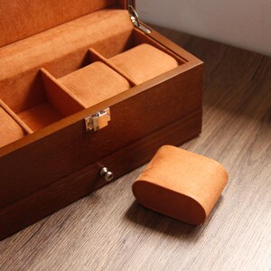 May include: A wooden watch box with a hinged lid, finished in a dark brown stain. The interior is lined with orange fabric and has compartments for watches. An orange watch pillow sits on the wooden surface, next to the box.