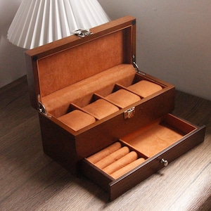May include: A wooden watch box, stained a dark brown, with a hinged lid and a pull-out drawer. The interior is lined with a soft, orange-brown material. The top section has compartments for watches, and the drawer has slots for accessories.