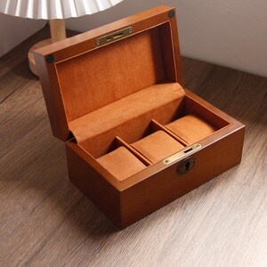 May include: A wooden watch box, open to show a soft, orange-brown interior. The box has three padded compartments for watches. The exterior is a warm, medium brown with a brass-coloured clasp and hinges. The box is likely to be around 20 cm in length.
