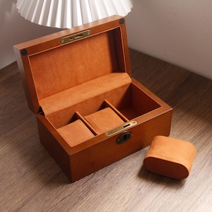 May include: A wooden watch box with a hinged lid, open to reveal a soft, orange interior with three watch pillows. The exterior is a warm brown with brass-coloured hardware. A separate watch pillow rests beside the box.