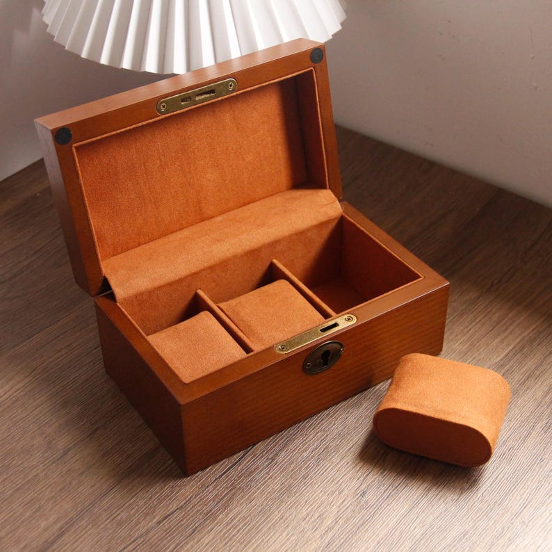 May include: A wooden watch box with a hinged lid, open to reveal a soft, orange interior with three watch pillows. The exterior is a warm brown with brass-coloured hardware. A separate watch pillow rests beside the box.