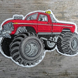 Könnte beinhalten: Gestickter Aufnäher eines roten Monstertrucks mit übergroßen grauen Reifen. Der Truck hat eine weiße Kontur und Details wie Auspuffrohre und eine gelbe Lichtleiste. Ein lustiges, verspieltes Design.