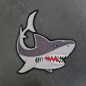 May include: Embroidered shark patch with a gray body, white accents, and a black outline. The shark's mouth is open, revealing red teeth. The patch is designed to be sewn onto clothing or accessories.