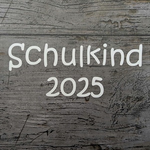 May include: White text on a gray wooden background reads "Schulkind 2025". The text is in a whimsical font. The background has a textured, weathered appearance, with visible wood grain.