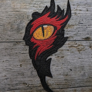 Könnte beinhalten: Gestickter Aufnäher mit einem Drachenauge. Das Auge hat eine gelbe Iris und eine schwarze Pupille, umrahmt von roten und schwarzen Stickdetails. Das Patch-Design enthält scharfe, eckige Elemente.