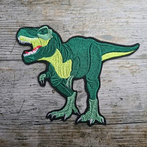 Könnte beinhalten: Gestickter Aufnäher mit einem grünen Tyrannosaurus Rex Dinosaurier mit gelben Akzenten auf Brust und Schwanz. Der Dinosaurier hat ein detailliertes Design mit offenem Maul, weißen Zähnen und roter Zunge. Der Aufnäher ist schwarz umrandet.