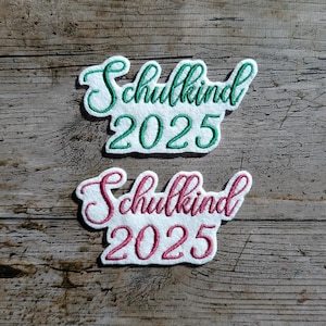 May include: Two embroidered patches with the text "Schulkind 2025". One patch has green lettering and the other has pink lettering, both on a white background. The patches are on a wooden surface.
