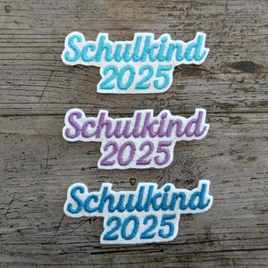 May include: Three embroidered patches with the text "Schulkind 2025" in a script font. Two patches are light blue with white trim, and one is purple with white trim. The patches are on a weathered wood surface.