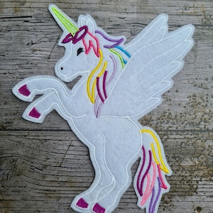 Peut inclure: Licorne en feutre blanc avec crinière, queue et corne arc-en-ciel. La licorne a de grandes ailes blanches et se tient sur ses pattes arrière. La crinière et la queue présentent des couleurs jaune, rose, violet et bleu. Les sabots de la licorne sont magenta.