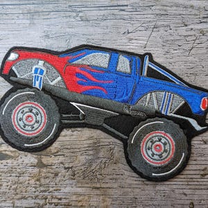 Könnte beinhalten: Gestickter Aufnäher eines Monster Trucks in Rot und Blau mit grauen Rädern und Akzenten. Der Truck hat Flammendesigns an der Seite und ist vor einem schwarzen Hintergrund platziert. Der Aufnäher ist für das Aufnähen auf Kleidung oder Accessoires gedacht.