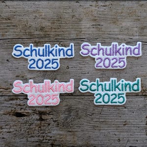 May include: Four embroidered patches with the German word "Schulkind" and the year "2025". The patches are in blue, purple, pink, and teal, with white outlines. The patches are on a wooden background.