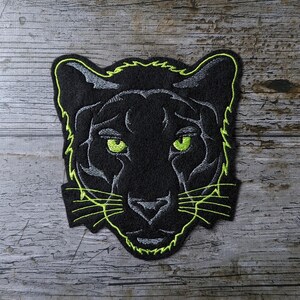 Könnte beinhalten: Gestickter schwarzer Panther-Aufnäher mit neongrünen und grauen Akzenten. Der Aufnäher zeigt ein Panthergesicht mit grünen Augen und einer grauen Nase. Die Ränder sind neongrün umrandet.