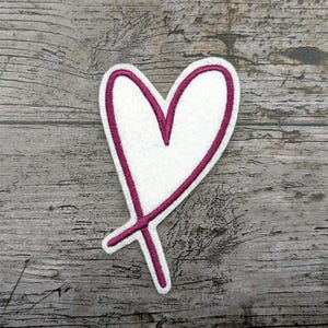 Könnte beinhalten: Ein weißes herzförmiges Aufnäher mit magentafarbenem Umriss und einem kleinen Kreuz am unteren Rand. Der Aufnäher befindet sich auf einer verwitterten, grauen Holzoberfläche. Dieser Aufnäher könnte zum Dekorieren von Kleidung oder Accessoires verwendet werden.